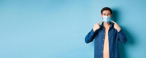 Coronavirus and pandemic concept. Portrait of happy and healthy man pointing fingers at his medical mask, smiling at camera, standing on blue background photo