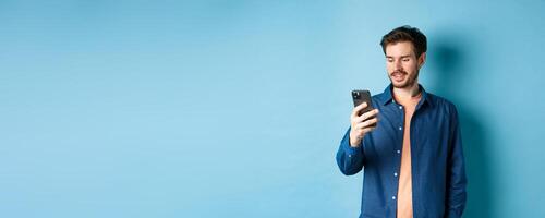 Handsome modern guy using mobile phone, reading smartphone screen and smiling, networking on blue background photo