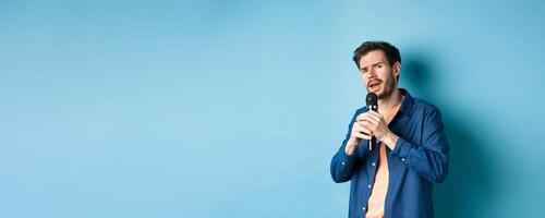 Young man singing song in microphone, playing karaoke, standing on blue background photo