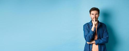 Modern guy shushing at camera, press finger to lips and telling a secret, making taboo sign, standing on blue background photo