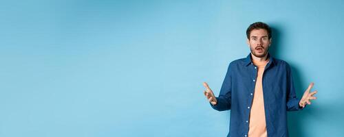 Shocked young man raising hands up and staring at camera startled, look with disbelief, standing on blue background photo