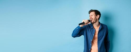 Carefree guy singing karaoke with microphone, perform a song, standing on blue background photo
