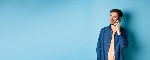 Handsome caucasian man talking on mobile phone, looking aside at empty space with carefree smile, standing on blue background photo
