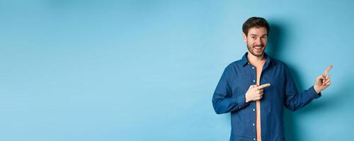 Cheerful caucasian man with beard pointing fingers aside at empty space and smiling at camera, showing advertisement, blue background photo