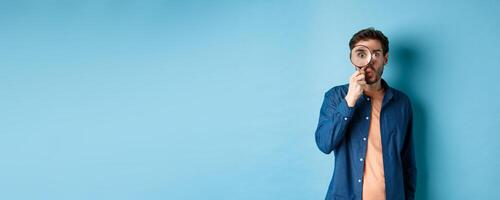 Image of surprised man found something interesting, say wow and look through magnifying glass, standing on blue background photo