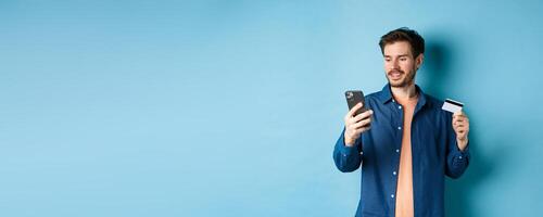 E-commerce concept. Image of handsome caucasian man paying online, looking at mobile phone screen and holding plastic credit card, blue background photo