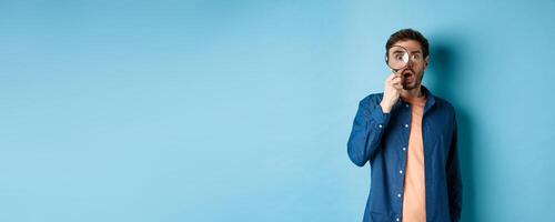 Impressed man look in awe through magnifying glass, drop jaw and stare at camera, standing on blue background photo