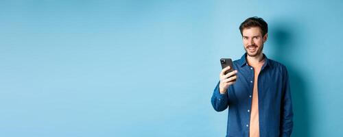 Attractive caucasian man using mobile phone, looking happy and smiling at camera, standing on blue background photo