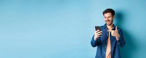 Image of handsome smiling man reading mobile screen and pointing at phone, checking out online promo or smartphone app, standing on blue background photo
