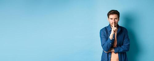 Angry man shushing, press finger to lips and frowning, tell to be quiet, show taboo gesture, standing on blue background photo