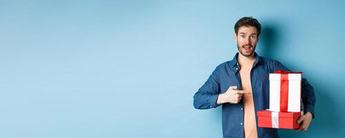 Handsome young man pointing at valentines gift boxes, standing over blue background in casual clothes photo
