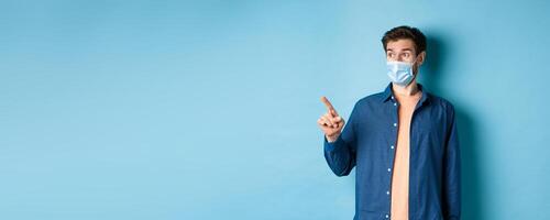 Covid-19 and healthcare concept. Surprised guy in face mask pointing and looking left at empty space, standing on blue background photo
