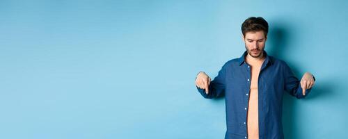 Image of modern guy in casual clothes, looking and pointing fingers down at empty space, reading logo, standing on blue background photo
