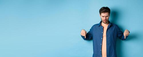 Confused and shocked guy looking and pointing down at something strange, standing on blue background photo