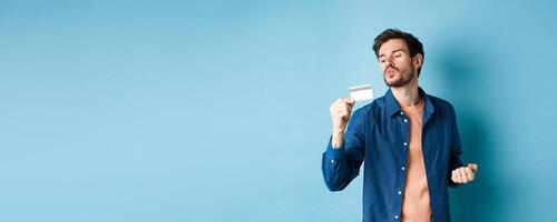 Funny guy kissing his plastic credit card and looking satisfied, shopping with discounts, standing on blue background photo