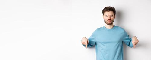 Sulking man pointing fingers down at something bad, frowning and complaining, standing over white background photo