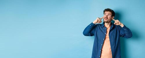 Annoyed young man block sound, shut ears with fingers and grimacing, disturbed by loud neighbours, looking up irritated, standing on blue background photo