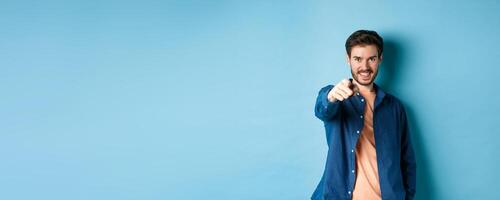 Confident smiling guy choosing or inviting you, pointing finger at camera decisive, standing on blue background photo
