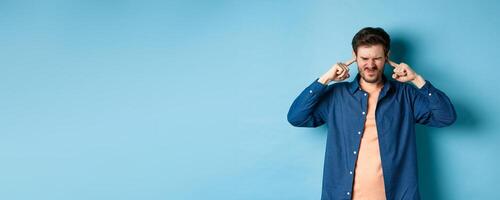 Annoyed young man block sound, shut ears with fingers and grimacing, disturbed by loud music, standing on blue background photo