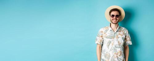 Handsome smiling guy in sunglasses and summer shirt, enjoying vacation on tour, standing on blue background photo