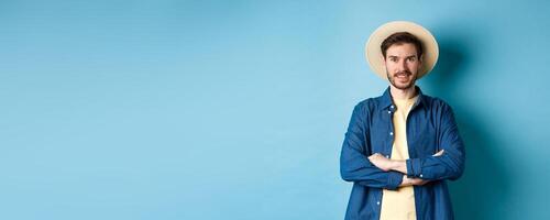 Portrait of cheerful guy going on vacation in straw summer hat, cross arms on chest and smiling, looking confident, standing on blue background photo