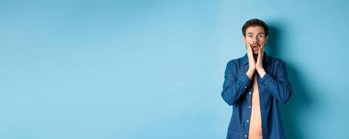 Portrait of guy gasping and dropping jaw with shocked face, staring at camera startled, standing on blue background photo