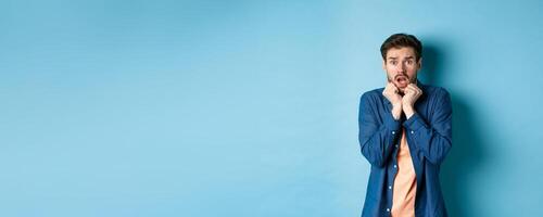 Scared young man gasping and screaming frightened, looking terrified at something, standing on blue background photo