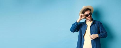 Handsome tourist in sunglasses and straw hat looking aside, smiling pleased, exploring sighsteeing on vacation, blue background photo