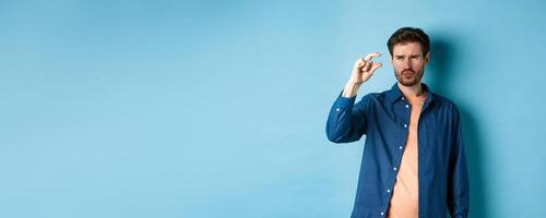 Confused guy showing small size and looking doubtful or skeptical, standing over blue background photo