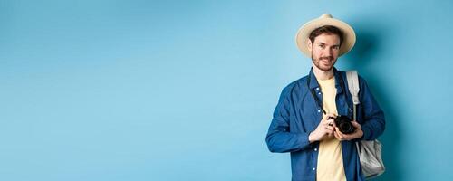 Handsome guy going on summer vacation, backpacking on holiday. Tourist with straw hat and camera smiling happy, standing on blue background photo