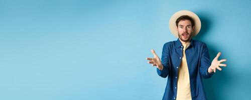 Surprised young man in summer hat, raising hands up staring with disbelief and amazement at camera, impressed with big event, standing on blue background photo