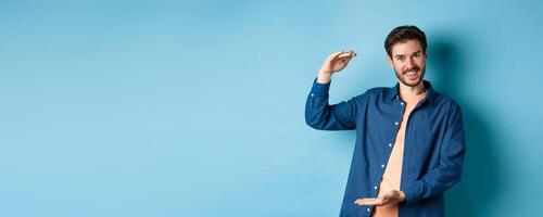 Handsome caucasian guy showing something big, shaping large size object with hands and smiling, standing on blue background photo