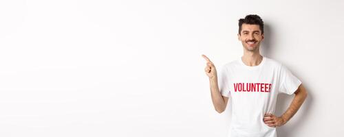 Handsome smiling volunteer in t-shirt pointing finger left, showing announcement, standing over white background photo