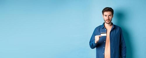 Handsome young man with beard, wearing casual clothes, showing plastic credit card and looking at camera, blue background photo