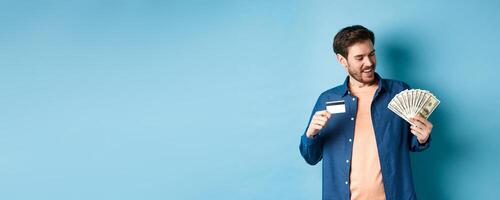 Smiling modern guy looking at cash and showing plastic credit card, standing on blue background photo