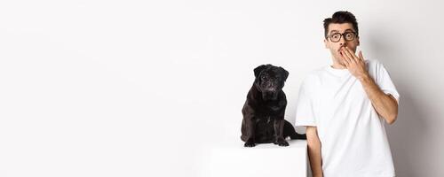 Image of dog owner and cute black pug looking at camera surprised and amazed, standing over white background photo