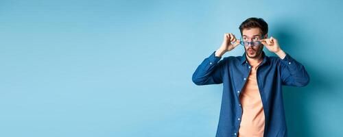 Image of caucasian guy takes-off sunglasses and looking aside with surprised face, checking out empty space for logo, standing on blue background photo