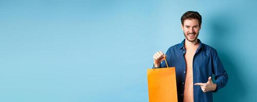 Smiling caucasian man pointing finger at orange shopping bag with bought items, standing on blue background photo