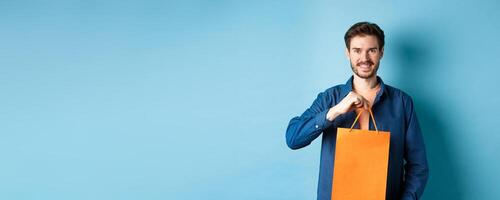 Handsome young man with beard, smiling and showing shopping bag, buying something in store, standing on blue background photo