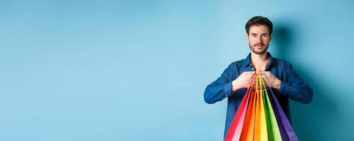 Modern guy with beard, holding packages from shop and smiling, standing on blue background photo