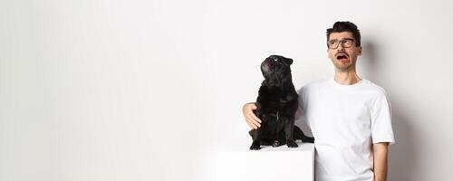 Whining man looking sad while pug staring intrigued at upper left corner, standing against white background photo