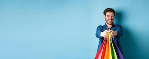 Cheerful handsome man winking and smiling, extending hands with colorful shopping bags, standing on blue background photo
