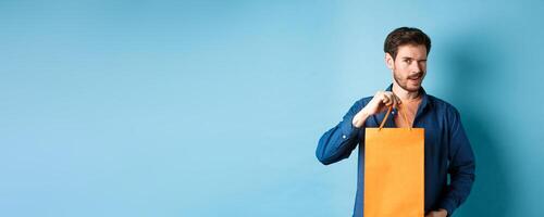 Happy young man showing orange shopping bag and winking, recommending shop discounts, standing on blue background photo