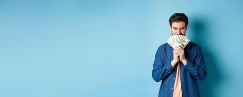 Young man hiding smiling face behind money and looking determined at camera, standing on blue background photo