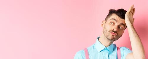 Close-up of handsome caucasian guy with moustache, feeling confident, touching his haircut and looking aside with smug face, standing on pink background photo