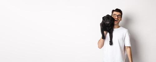 Cheerful hipster guy in glasses and t-shirt, carry cute black pug on shoulder and smiling. Dog owner playing with his pet, standing over white background photo