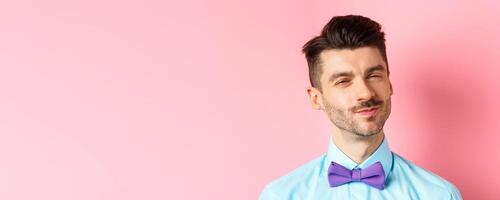 Close-up of smiling young guy with moustache, squinting intrigued, listening to something interesting, standing in bow-tie on pink background photo