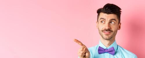Close-up of caucasian male model with moustache and bow-tie, looking and pointing left with intrigued smile, showing promo offer, standing in bow-tie on pink background photo