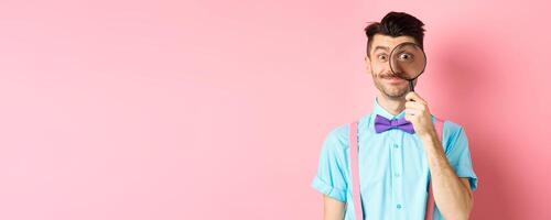 Funny caucasian man with moustache and bow-tie, looking through magnifying glass with big eye and smiling, standing over pink background photo
