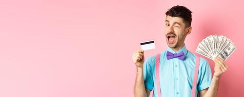 Cheerful caucasian man in bow-tie winking at you, showing plastic credit card and money, recommending special deal, standing on pink background photo